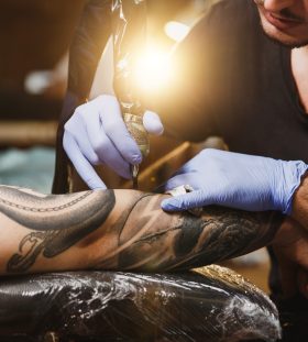 Close up of professional tattooer artist doing picture on hand of man by machine black ink from a jar. Tattoo art on body. Equipment for making tattoo art. Master makes tattooed in light studio.